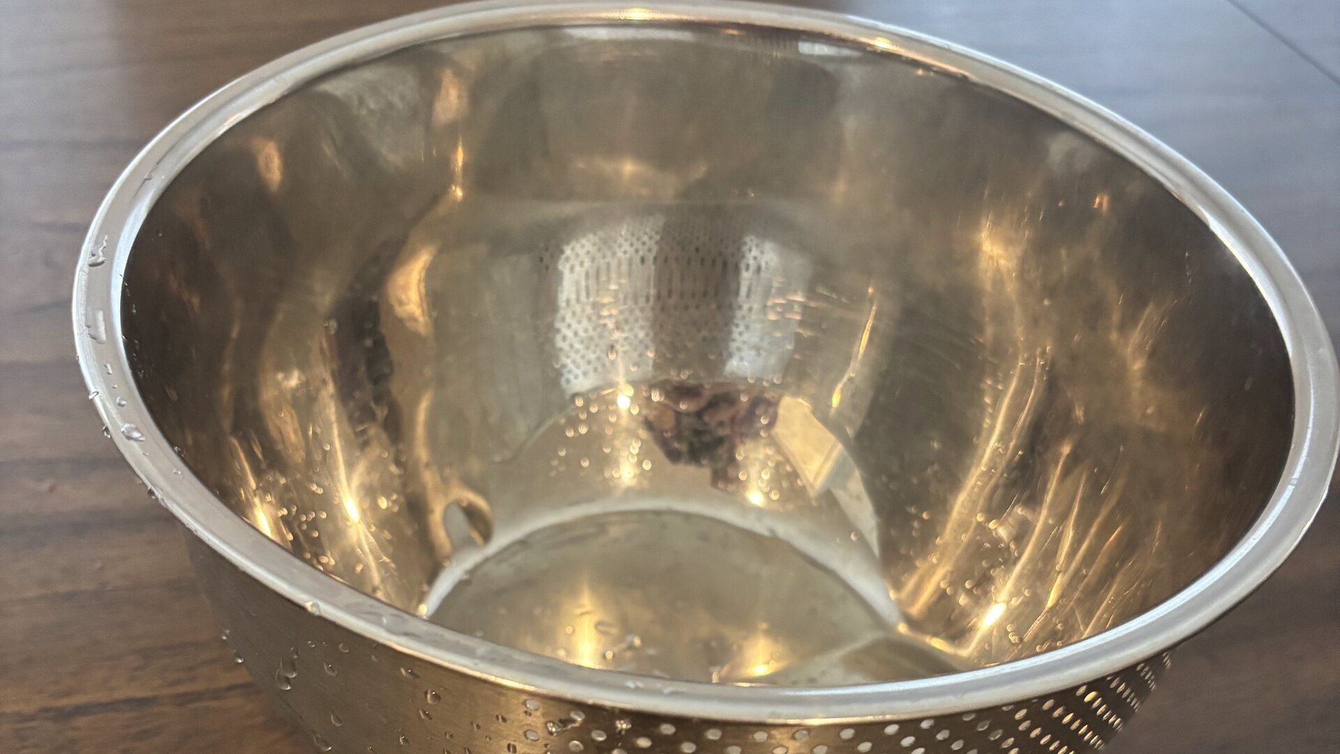 A stainless rice-washing bowl on a wooden table, its lower sides pierced with drain holes