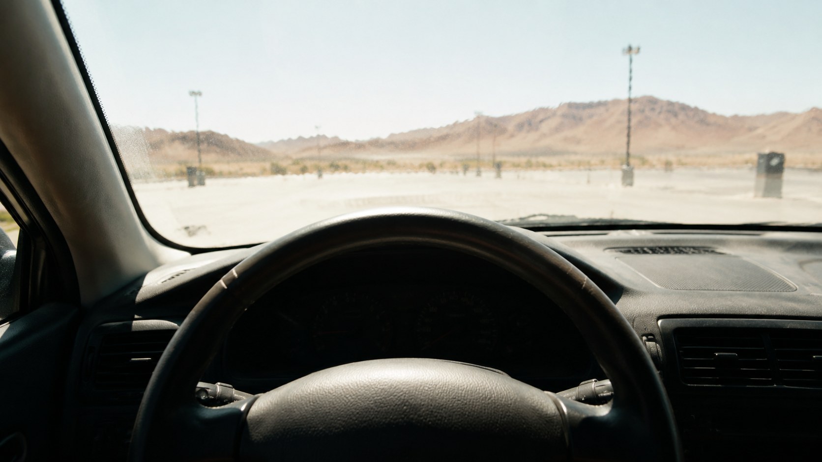The driver's seat of a car parked in the desert sun, looking over a dark steering wheel and dashboard to a bleached empty lot and dry hills (illustration)