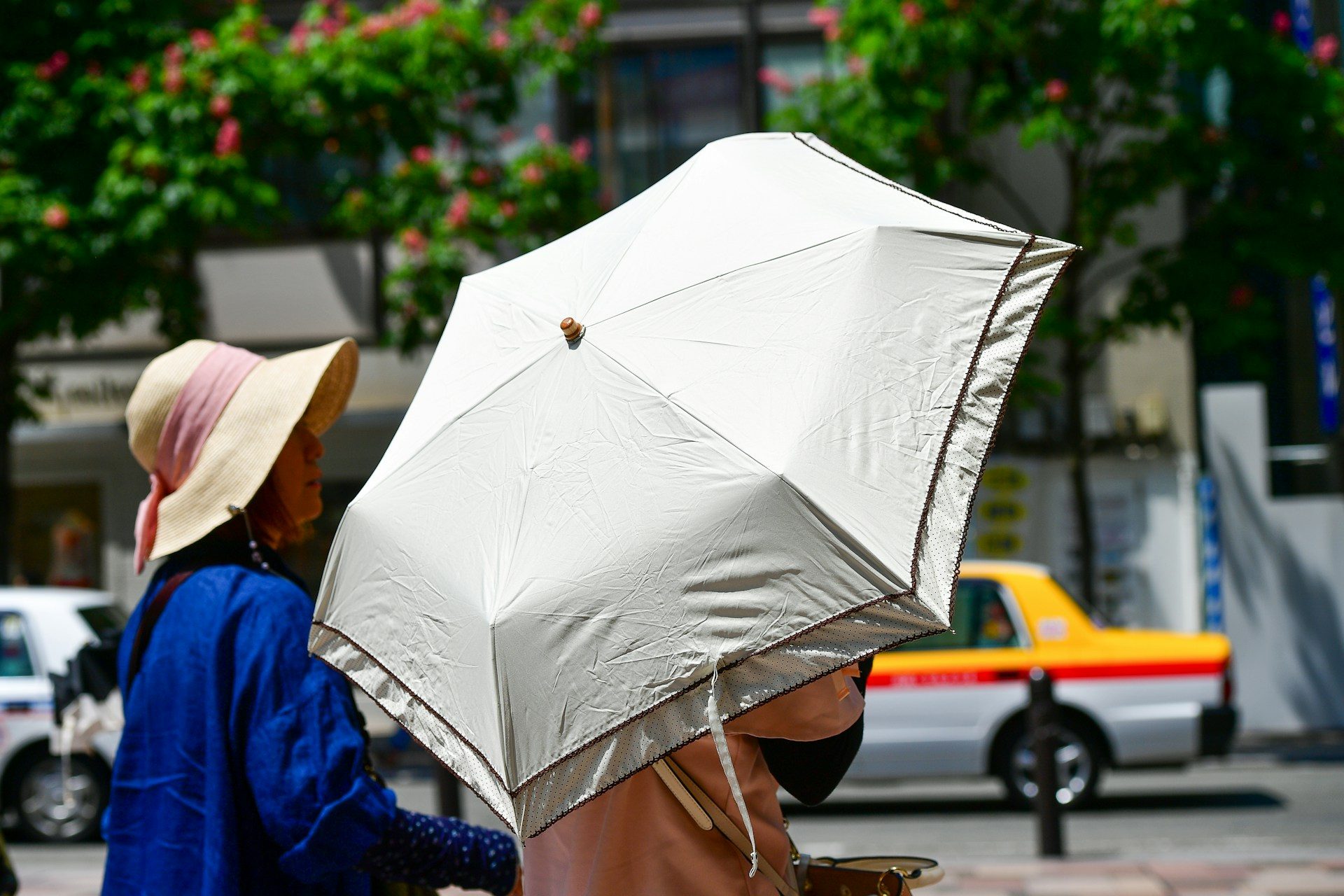 A woman walking a Japanese city street in summer under an open sun umbrella, a taxi passing behind her
